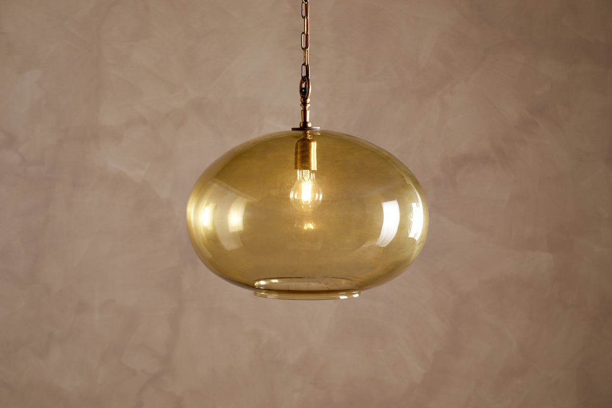The Otoro Recycled Glass Pendant - Honey - Large Round by nkuku hangs from the ceiling, featuring a round, amber-tinted recycled glass shade with brass fittings and a chain. An exposed bulb glows softly inside, casting warm light against a textured, light beige wall.