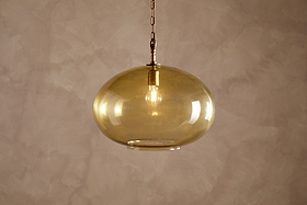 The Otoro Recycled Glass Pendant - Honey - Large Round by nkuku hangs from the ceiling, featuring a round, amber-tinted recycled glass shade with brass fittings and a chain. An exposed bulb glows softly inside, casting warm light against a textured, light beige wall.