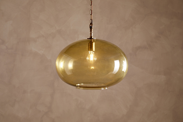 The Otoro Recycled Glass Pendant - Honey - Large Round by nkuku hangs from the ceiling, featuring a round, amber-tinted recycled glass shade with brass fittings and a chain. An exposed bulb glows softly inside, casting warm light against a textured, light beige wall.