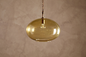 The Otoro Recycled Glass Pendant - Honey - Large Round by nkuku features a round, amber-toned shade crafted from recycled glass, suspended from a gold chain with refined brass fittings against a beige textured wall. The clear glass showcases an exposed bulb within the fixture, offering a contemporary and minimalist aesthetic.