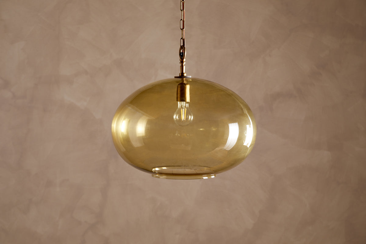 The Otoro Recycled Glass Pendant - Honey - Large Round by nkuku features a round, amber-toned shade crafted from recycled glass, suspended from a gold chain with refined brass fittings against a beige textured wall. The clear glass showcases an exposed bulb within the fixture, offering a contemporary and minimalist aesthetic.