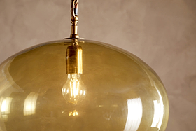 A close-up of the Otoro Recycled Glass Pendant - Honey - Large Round by nkuku, showcasing a glowing bulb within its round, translucent amber glass shade and elegant brass fittings. The pendant hangs from a wire, with soft reflections visible on the recycled glass surface. The softly textured beige background enhances its warm, inviting glow.