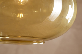 Here’s a rewritten version incorporating the product data: A close-up of the Otoro Recycled Glass Pendant in Honey (Large Round) by nkuku shows off its translucent amber glass shade with gently curved surfaces, reflecting light and hints of a window. Subtle brass fittings add an elegant accent, while the blurred beige background highlights the unique recycled glass texture and inviting warm glow within the pendant.