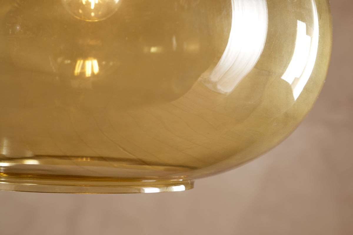 Here’s a rewritten version incorporating the product data: A close-up of the Otoro Recycled Glass Pendant in Honey (Large Round) by nkuku shows off its translucent amber glass shade with gently curved surfaces, reflecting light and hints of a window. Subtle brass fittings add an elegant accent, while the blurred beige background highlights the unique recycled glass texture and inviting warm glow within the pendant.