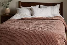A neatly made bed with white pillows, a striped white sheet, and the nkuku Nidra Velvet & Linen Bed Quilt in Rust. A wooden headboard and a nightstand with greenery complete this King Super King bedding setup.