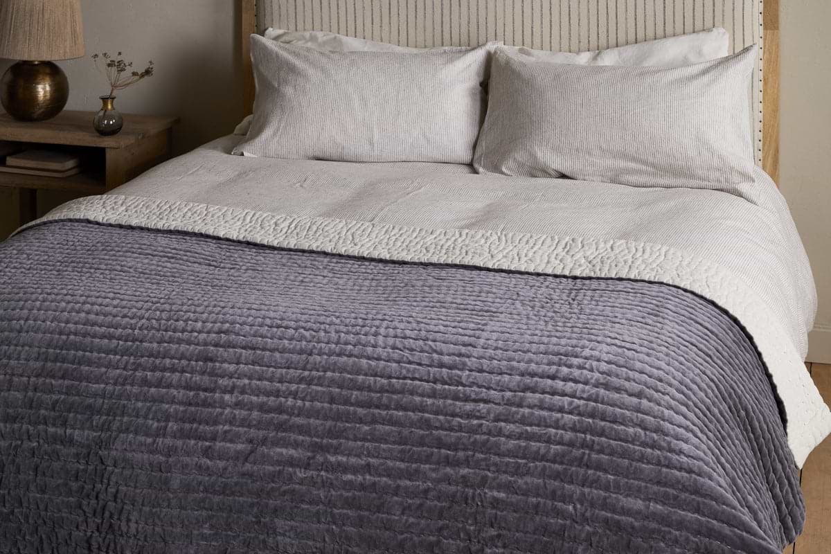 The nkuku Nidra Velvet & Linen Bed Quilt in Charcoal, with Kantha quilting, is styled on a neatly made bed with white pillows and a beige blanket. A wooden nightstand with a vase and lamp sits beside—ideal for King and Super King beds.