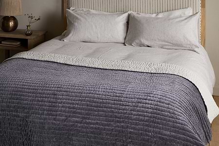 The nkuku Nidra Velvet & Linen Bed Quilt in Charcoal, with Kantha quilting, is styled on a neatly made bed with white pillows and a beige blanket. A wooden nightstand with a vase and lamp sits beside—ideal for King and Super King beds.