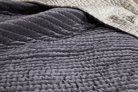 A close-up of the textured Nidra Velvet & Linen Bed Quilt in charcoal by nkuku, layered over a ribbed gray blanket. The soft, cozy Kantha-quilted fabric is ideal for King and Super King beds.