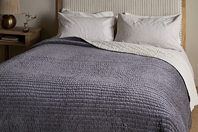 A neatly made bed features the nkuku Nidra Velvet & Linen Bed Quilt in Charcoal, layered over a white blanket with two large white pillows and a beige headboard. Ideal for King and Super King beds, with a wooden nightstand, vase, and lamp beside it.