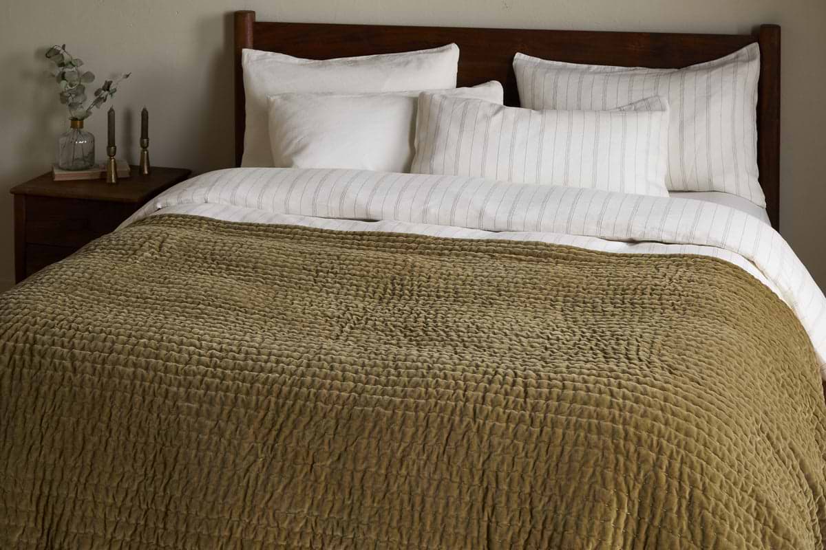 A king size bed is dressed with white pillows, a cream-striped blanket, and the nkuku Nidra Velvet & Linen Bed Quilt in Moss, featuring Kantha quilting. A wooden headboard and nightstand with decor items are in the background.