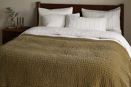 A king size bed is dressed with white pillows, a cream-striped blanket, and the nkuku Nidra Velvet & Linen Bed Quilt in Moss, featuring Kantha quilting. A wooden headboard and nightstand with decor items are in the background.