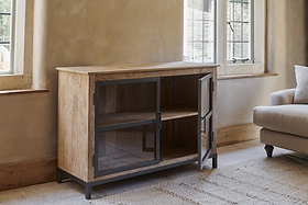 The Nirvi Mango Wood & Iron Sideboard - Black by nkuku, featuring black metal-framed glass doors, stands beside a window in a bright room. One door is open to show a shelf inside; a light armchair and rug are partly visible.