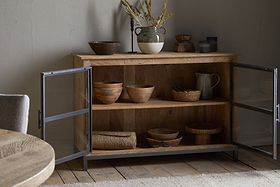 The nkuku Nirvi Mango Wood & Iron Sideboard - Black features black metal-framed glass doors that open to display wooden bowls, a pitcher, and baskets inside, while pottery and dried grasses decorate the top.