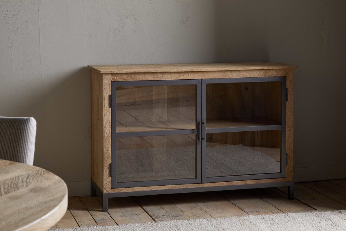 The nkuku Nirvi Mango Wood & Iron Sideboard - Black features glass-front double doors and a black frame, standing on a wooden floor against a light wall in a minimalist room with part of a round table and upholstered chair visible on the left.