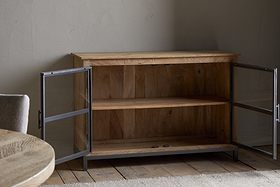 The Nirvi Mango Wood & Iron Sideboard by nkuku, featuring glass doors and a single shelf, sits open on a wooden floor beside a round table and cushioned chair in a softly lit room with textured walls.