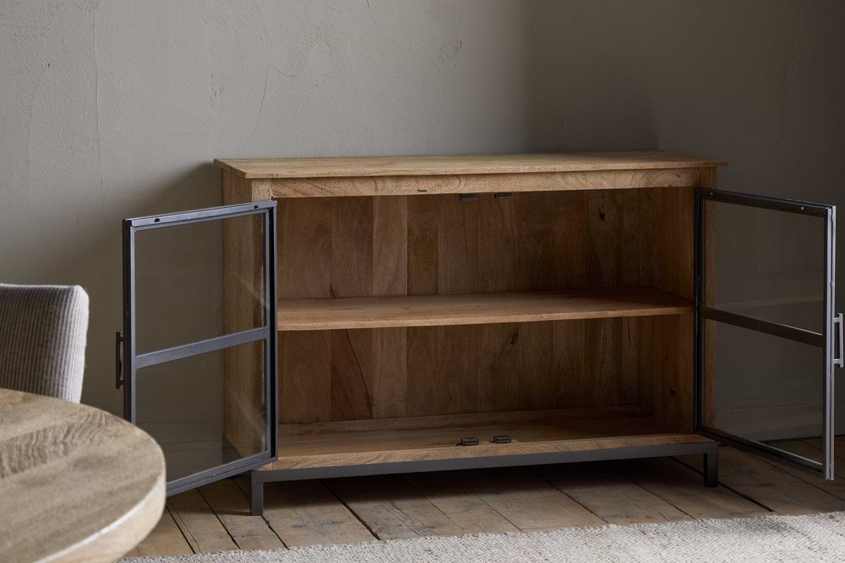 The Nirvi Mango Wood & Iron Sideboard by nkuku, featuring glass doors and a single shelf, sits open on a wooden floor beside a round table and cushioned chair in a softly lit room with textured walls.