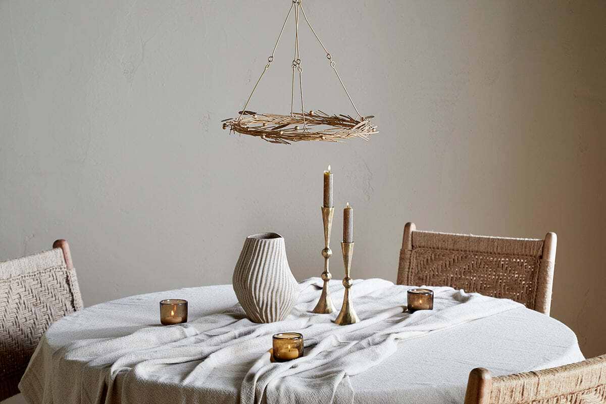 A round table with a cream cloth features a ribbed ceramic vase, brass candle holders, and brown votives. Two woven chairs sit nearby, all beneath the elegant nkuku Natesa Hanging Wreath - Brass and a rustic twig chandelier overhead.