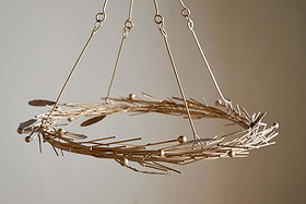 The Natesa Hanging Wreath - Brass by nkuku is a modern chandelier featuring thin gold rods, small spheres, and feather-shaped pieces. Suspended with four metal rods, it stands out against a plain beige background.