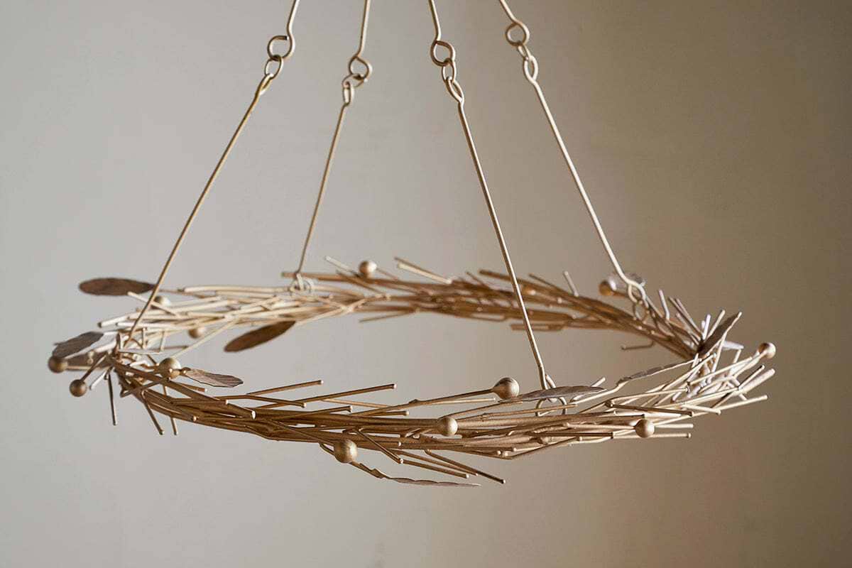 The Natesa Hanging Wreath - Brass by nkuku is a modern chandelier featuring thin gold rods, small spheres, and feather-shaped pieces. Suspended with four metal rods, it stands out against a plain beige background.