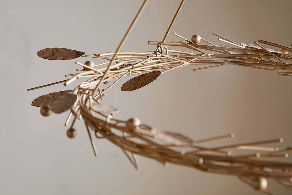 A close-up of the Natesa Hanging Wreath - Brass by nkuku showcases an abstract, circular sculpture crafted from thin brass rods, oval shapes, and small spheres. Its intertwined form evokes elegant traditional craftsmanship against a softly blurred background.