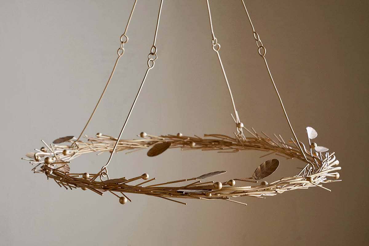 The Natesa Hanging Wreath - Brass by nkuku features delicate, light-hued twigs and brass leaves suspended by fine wires, highlighting traditional artistry and casting gentle shadows on any neutral wall.