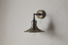 The Najja Brass Wall Lamp by nkuku features a vintage-style wide, shallow shade and exposed bulb in an antique brass finish, elegantly mounted on a light-colored, textured wall.
