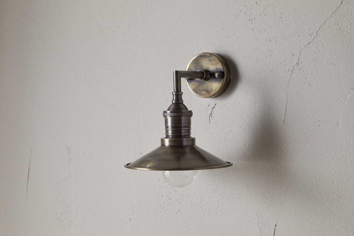 The Najja Brass Wall Lamp by nkuku features a vintage-style wide, shallow shade and exposed bulb in an antique brass finish, elegantly mounted on a light-colored, textured wall.