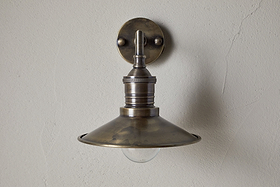 The Najja Brass Wall Lamp by nkuku features a vintage-style, wide shallow metal shade and exposed bulb in an antique brass finish, mounted on a light-colored textured wall.