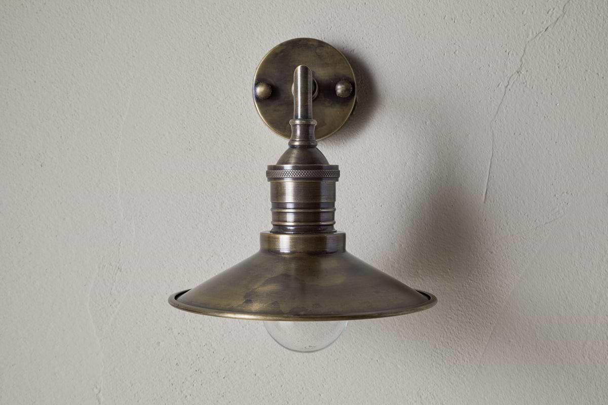 The Najja Brass Wall Lamp by nkuku features a vintage-style, wide shallow metal shade and exposed bulb in an antique brass finish, mounted on a light-colored textured wall.