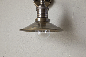 A close-up of the nkuku Najja Brass Wall Lamp in antique brass, featuring a metal shade and exposed clear bulb, mounted on a light-colored textured wall.