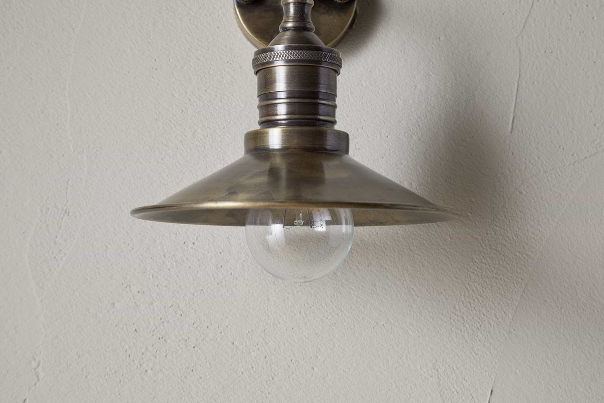 A close-up of the nkuku Najja Brass Wall Lamp in antique brass, featuring a metal shade and exposed clear bulb, mounted on a light-colored textured wall.
