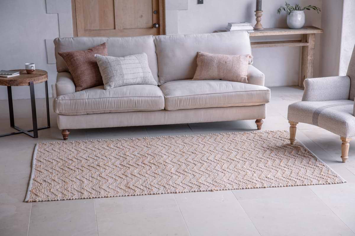 A neutral-toned living room features a beige sofa with four throw pillows, a Nabami Chevron Jute Rug - Natural by nkuku in cream and tan, a small round side table with a cup, wooden console with decor, potted plant, and light upholstered chair for a bright, cozy feel.