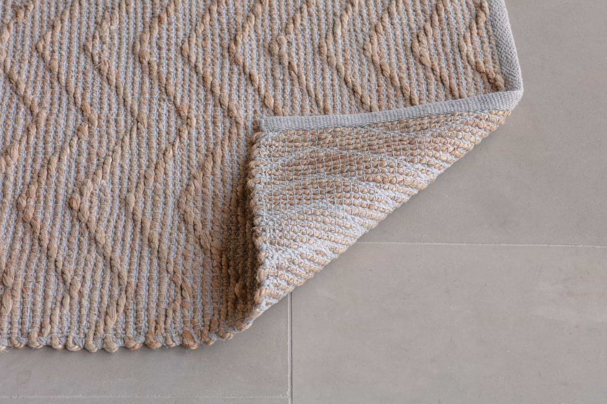 The Nabami Chevron Jute Rug - Natural by nkuku features a subtle chevron pattern in beige and light brown. Its stylish, textured design is shown on a light gray tiled floor with one corner folded back to display its thickness and detailed weave.