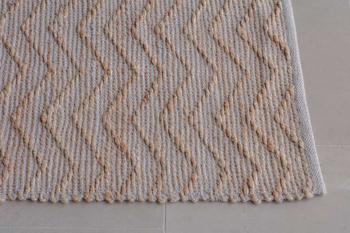 A close-up of the Nabami Chevron Jute Rug by nkuku shows its beige and light brown zigzag pattern and finished edge on a light gray tiled floor. The textured, braided jute design highlights its natural look.