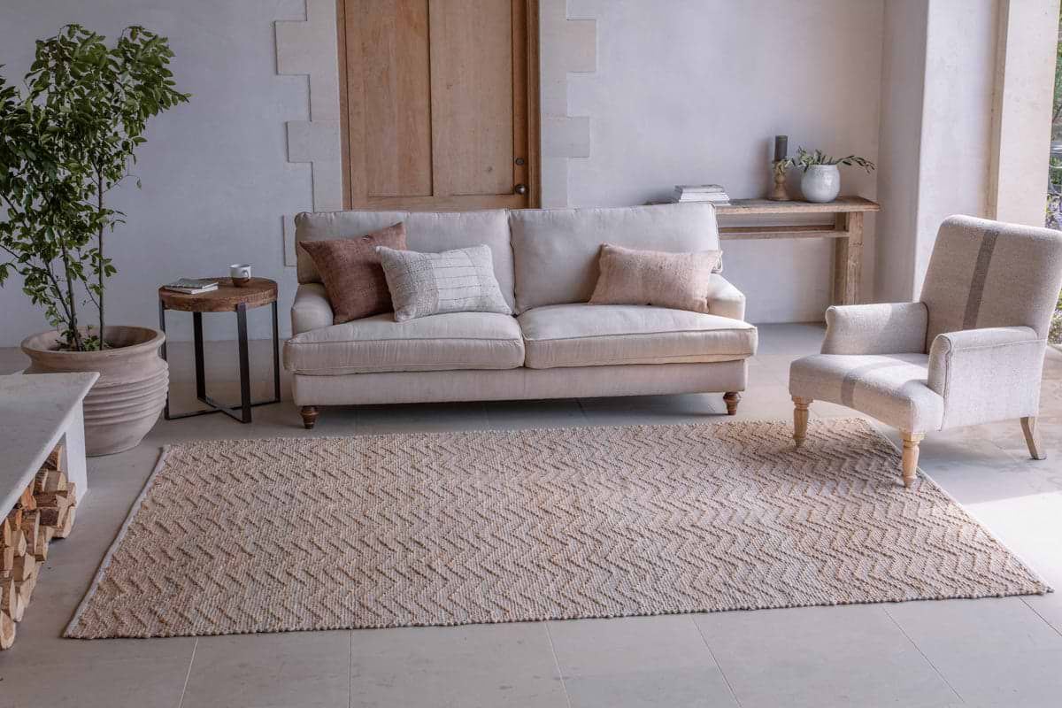A modern living room with light beige walls, a cream sofa, assorted cushions, a patterned armchair, a round side table with a plant, and the Nabami Chevron Jute Rug - Natural by nkuku as the large neutral rug, plus a wooden console table behind.