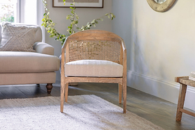 The Nabhi Mango Wood & Cane Occasional Chair by nkuku, featuring a woven rattan backrest and beige cushion, sits on a light rug. A beige sofa, wooden stool with books, and green leafy plants warm this sunlit living room.