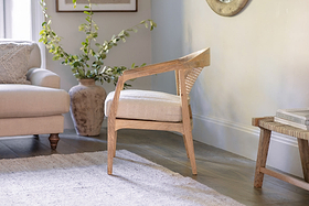 The nkuku Nabhi Mango Wood & Cane Occasional Chair features an upholstered cotton cushion and sits on a light rug. A beige sofa with a textured pillow is to the left, and a large textured vase with greenery stands behind the sofa.