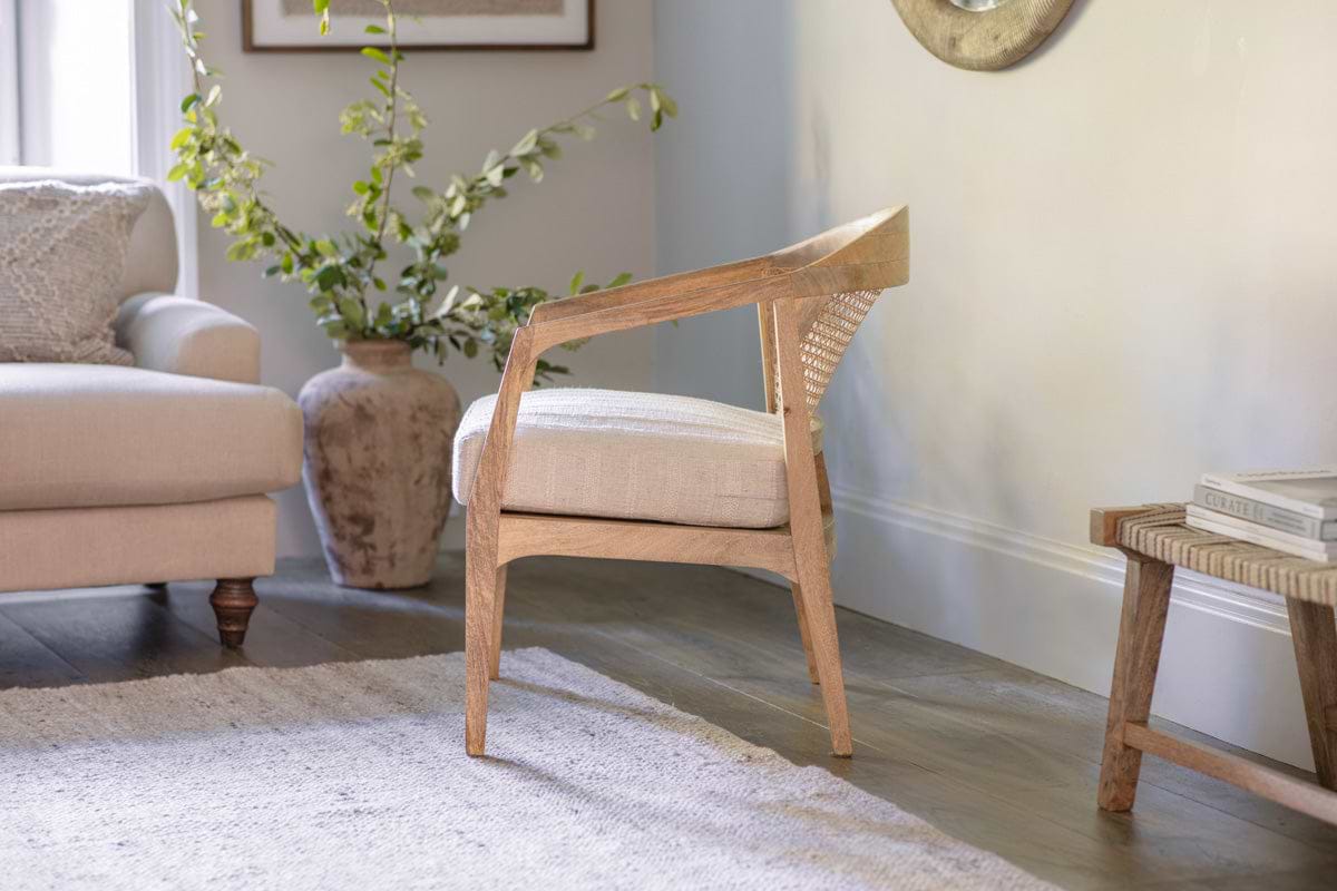 The nkuku Nabhi Mango Wood & Cane Occasional Chair features an upholstered cotton cushion and sits on a light rug. A beige sofa with a textured pillow is to the left, and a large textured vase with greenery stands behind the sofa.