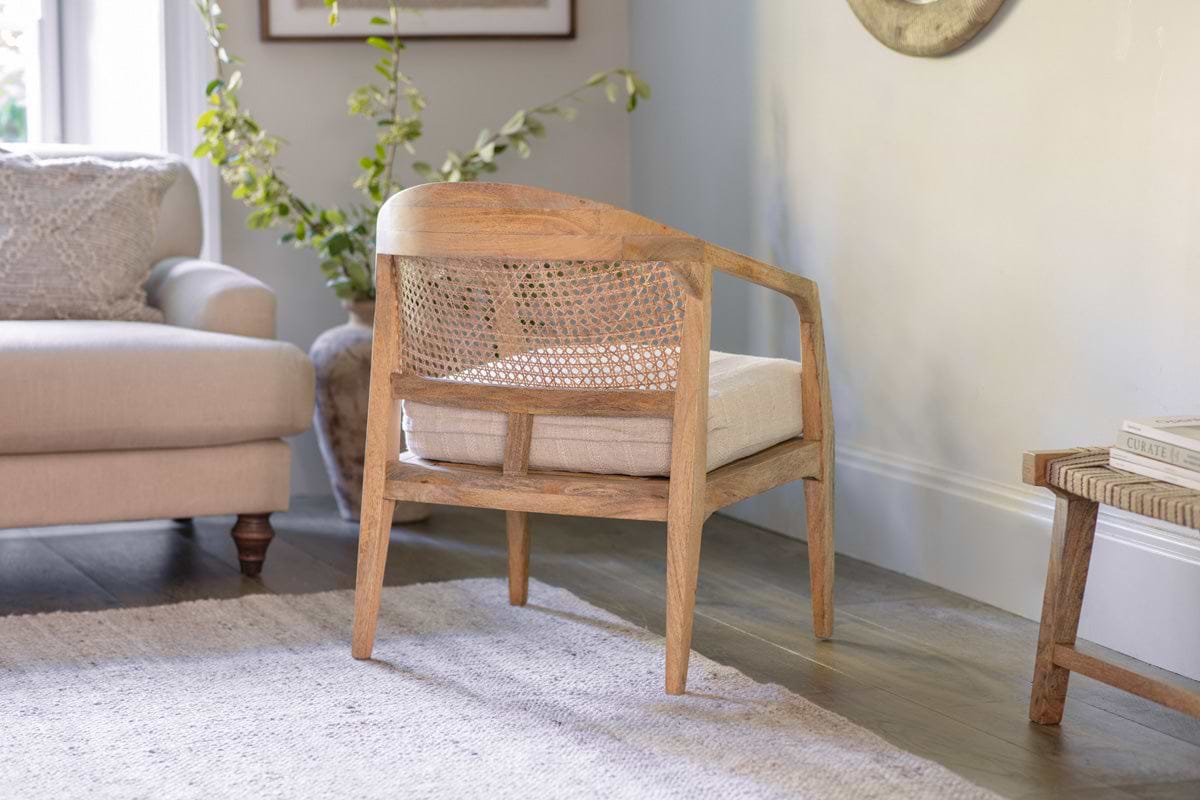 The Nabhi Mango Wood & Cane Occasional Chair by nkuku, featuring a natural mango wood frame and cane back with an upholstered cotton cushion, is shown from behind in a bright living room with a beige sofa, green plant, rug, and side table.