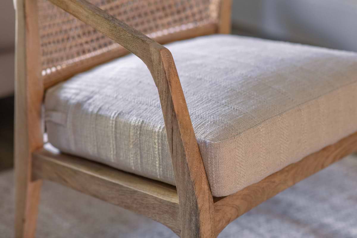 Close-up of the Nabhi Mango Wood & Cane Occasional Chair by nkuku, featuring a natural wood frame, handcrafted cane back, and thick cream linen cushion. Curved armrest showcases its mid-century modern elegance.