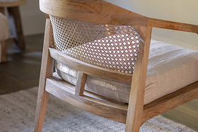 A close-up of the nkuku Nabhi Mango Wood & Cane Occasional Chair—Natural shows its curved cane backrest and woven mesh. Crafted from light mango wood with a plush cotton cushion, it sits on a rug and hardwood floor in the background.