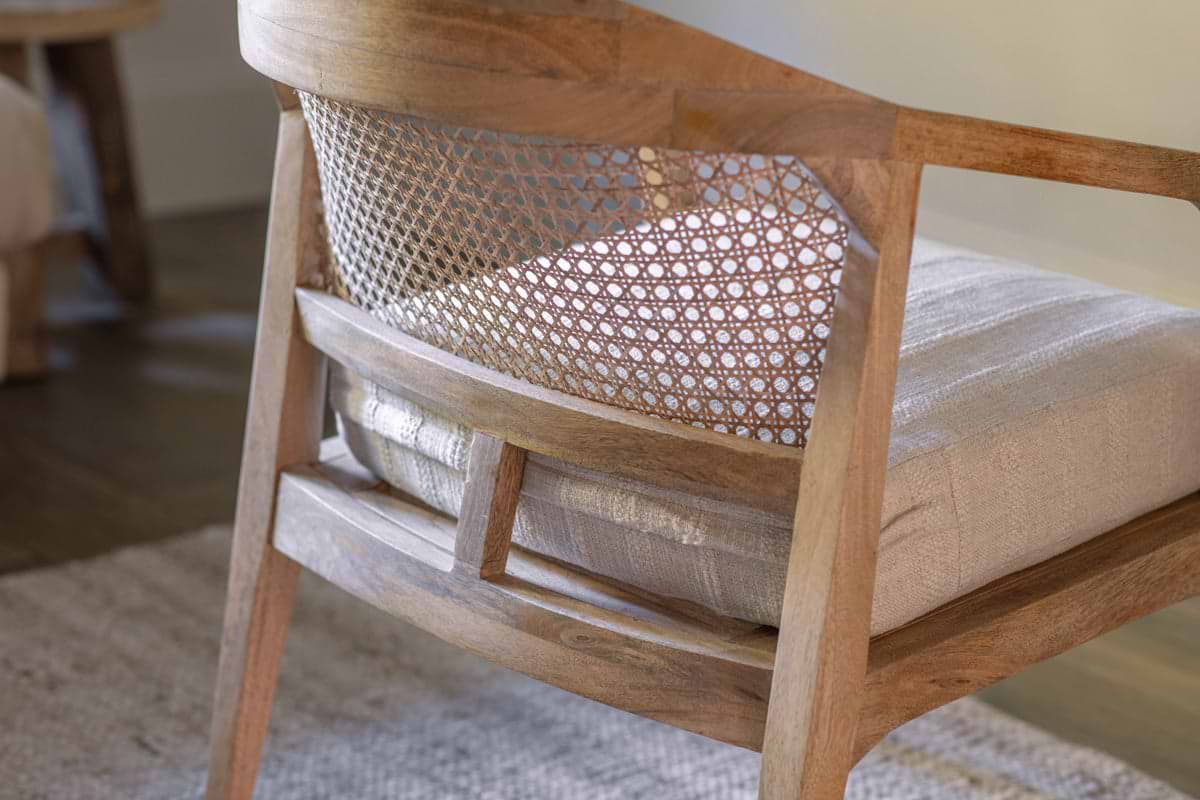 A close-up of the nkuku Nabhi Mango Wood & Cane Occasional Chair—Natural shows its curved cane backrest and woven mesh. Crafted from light mango wood with a plush cotton cushion, it sits on a rug and hardwood floor in the background.