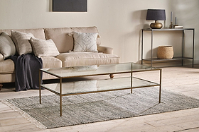 A modern living room with a beige sofa, neutral throw pillows, and the nkuku Nakuru Iron & Glass Coffee Table on a gray rug. A console table with decor and a woven basket stands against a light wall, all atop light wood flooring.
