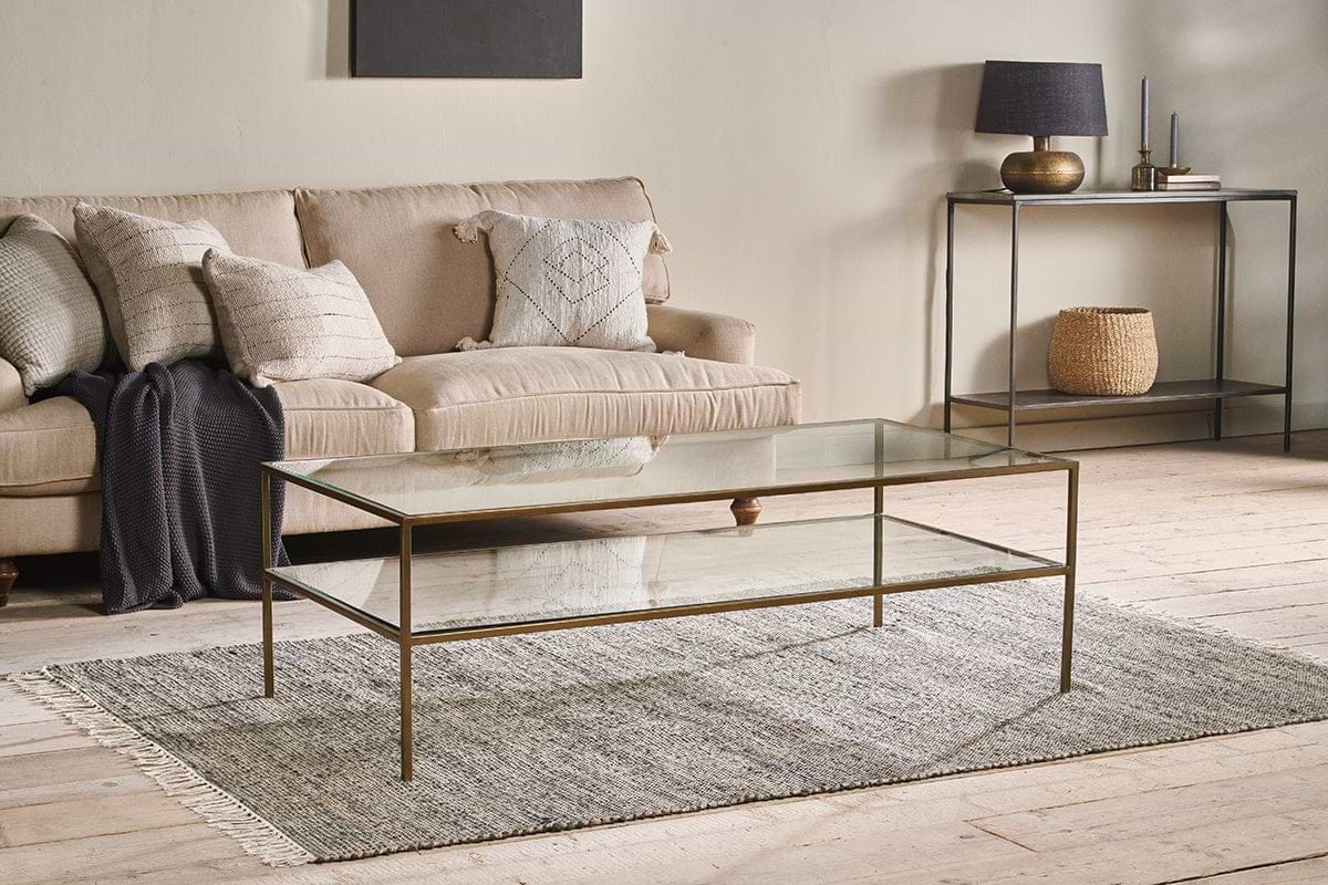 A modern living room with a beige sofa, neutral throw pillows, and the nkuku Nakuru Iron & Glass Coffee Table on a gray rug. A console table with decor and a woven basket stands against a light wall, all atop light wood flooring.