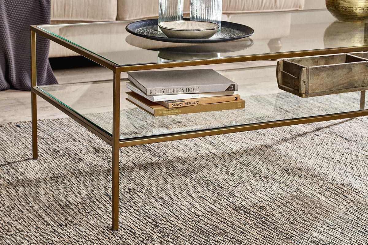 A nkuku Nakuru Iron & Glass Coffee Table with a gold frame stands on a textured gray rug. Its glass shelf holds books and a wooden box, while the tabletop displays a round tray, glass vase, and decor. A beige sofa is in the background.