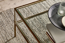 A nkuku Nakuru Iron & Glass Coffee Table stands on a textured woven carpet. On the table are stacked books, a dark patterned plate, and a ribbed glass tumbler. A wooden floor is visible in the upper left corner.