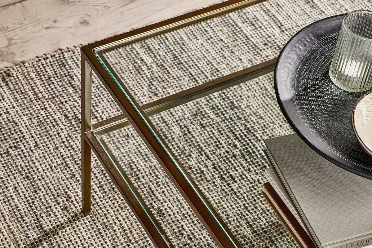 A nkuku Nakuru Iron & Glass Coffee Table stands on a textured woven carpet. On the table are stacked books, a dark patterned plate, and a ribbed glass tumbler. A wooden floor is visible in the upper left corner.