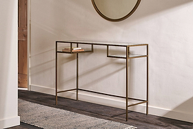 The Nkuku Nakuru Iron & Glass Console Table stands against a white wall, displaying two stacked books. A round mirror hangs above, while natural light from the left highlights the dark wood floor and textured rug.