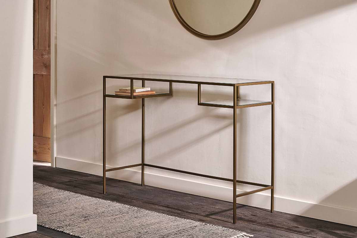 The Nkuku Nakuru Iron & Glass Console Table stands against a white wall, displaying two stacked books. A round mirror hangs above, while natural light from the left highlights the dark wood floor and textured rug.