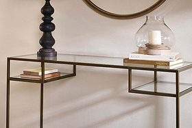 The nkuku Nakuru Iron & Glass Console Table, topped with stacked books, a black lamp, and a glass hurricane vase with a white candle, sits beneath a round mirror that reflects the table’s elegant, contemporary style.